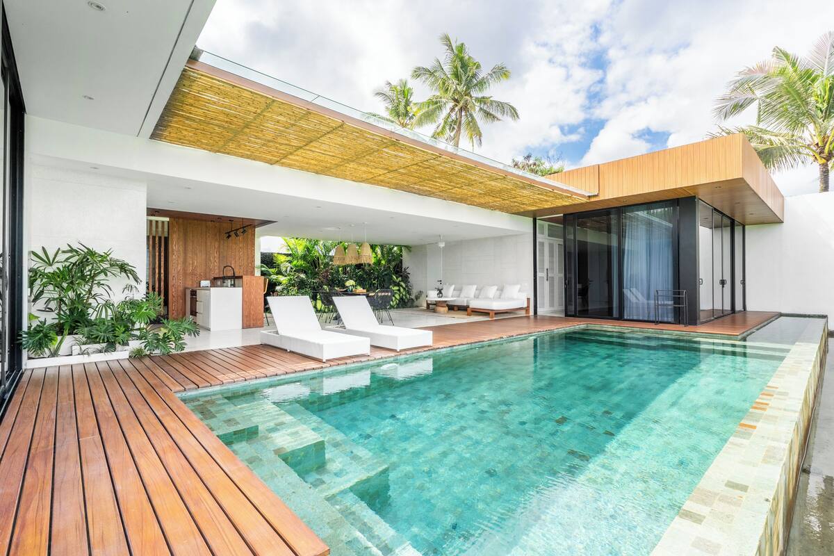A modern poolside area with a wooden deck, white lounge chairs, and a glass-enclosed space.