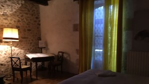 A dimly lit room with a stone wall, a small round table with chairs, a bed with a pillow, and a window with yellow curtains. - Charming cottage in Périgord: Aliénor's relay (Capdrot)