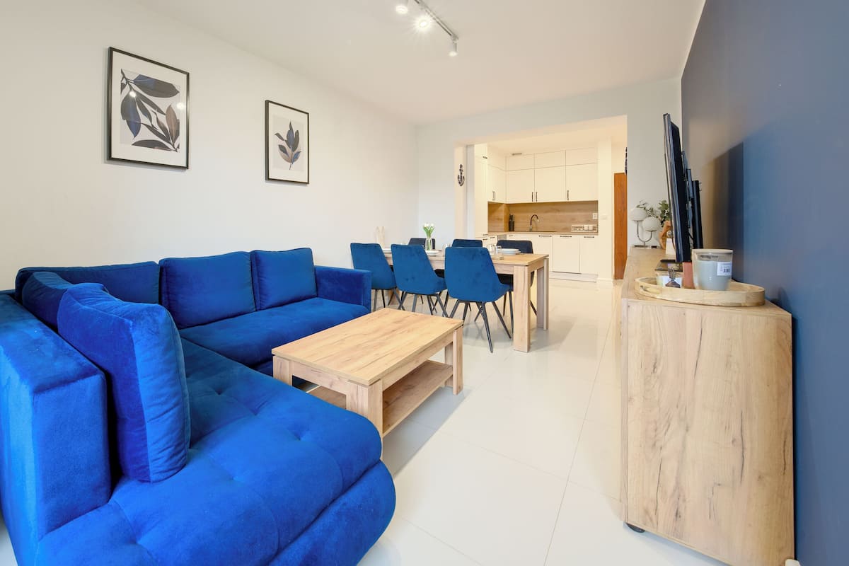 Un salon moderne avec un canapé bleu, une table basse en bois et un coin repas comprenant des chaises et une table. Des tableaux encadrés ornent le mur, et on aperçoit un coin cuisine à l’arrière-plan.