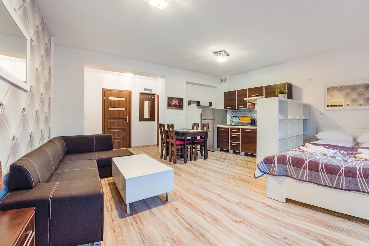 Une pièce moderne et bien éclairée, comprenant un canapé, une petite table, un lit avec une couverture à motifs, un coin cuisine avec des placards et un réfrigérateur, ainsi qu’une table à manger avec des chaises.