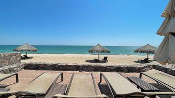Un patio frente a la playa con sillones reclinables y sombrillas de paja, con vista al mar.