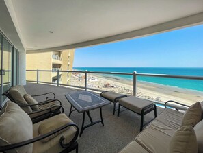 Varanda com área de estar externa, vista para a praia e um edifício ao fundo.