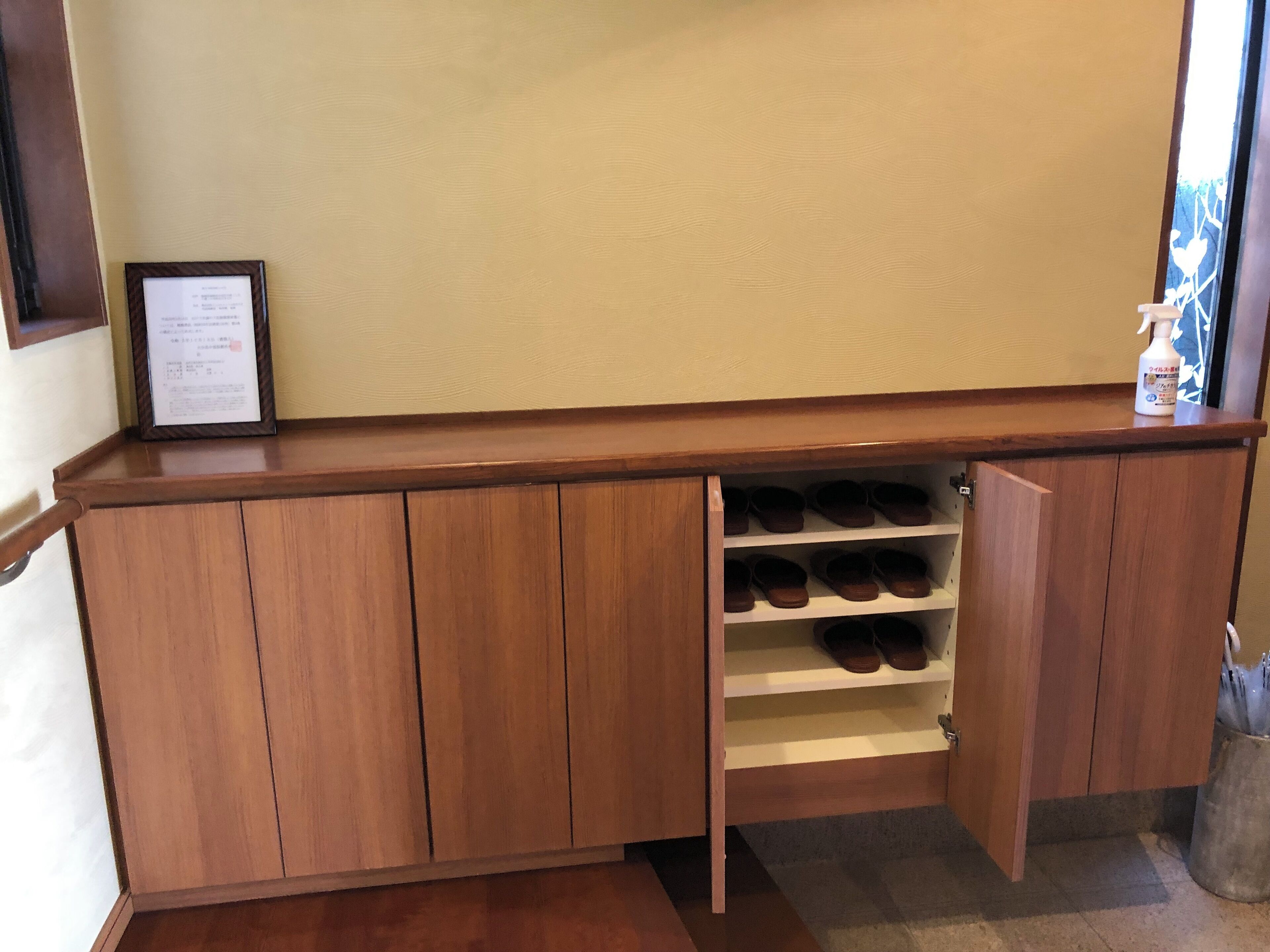 Een houten kast met meerdere schoenenplanken, een ingelijst certificaat bovenop en een fles handdesinfecterend middel aan de rechterkant.