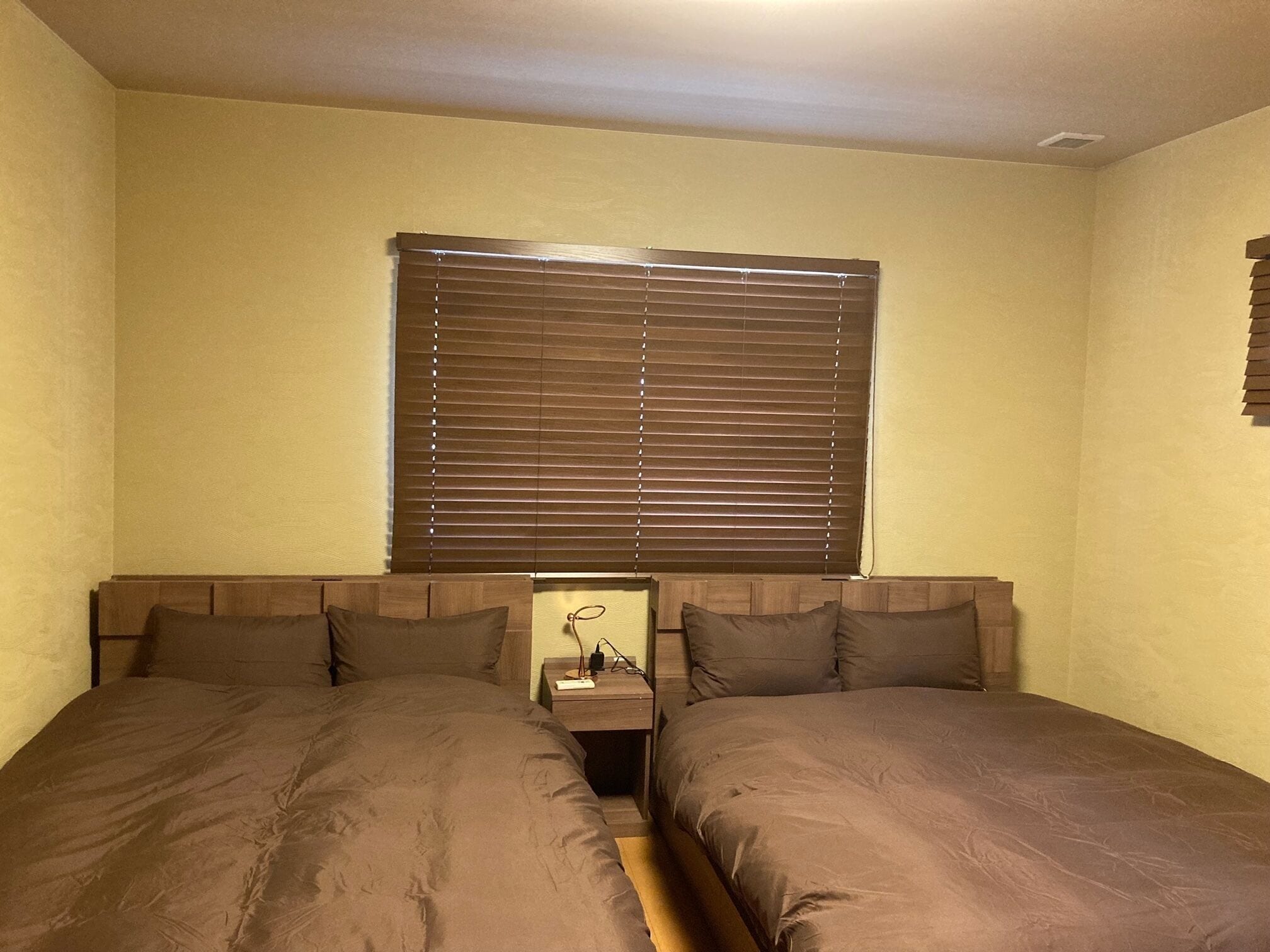 A bedroom with two single beds, a nightstand with a lamp, and a window with blinds.