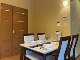 Un comedor con una mesa dispuesta para dos personas, equipada con platos, tazones, cubiertos y copas de vino. Hay dos sillas, una cafetera y un termostato en la pared.