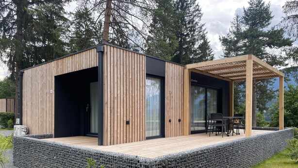 A modern wooden cabin with a covered patio, large windows, and a stone retaining wall. - EuroParcs Pressegger See (Hermagor-Pressegger See)