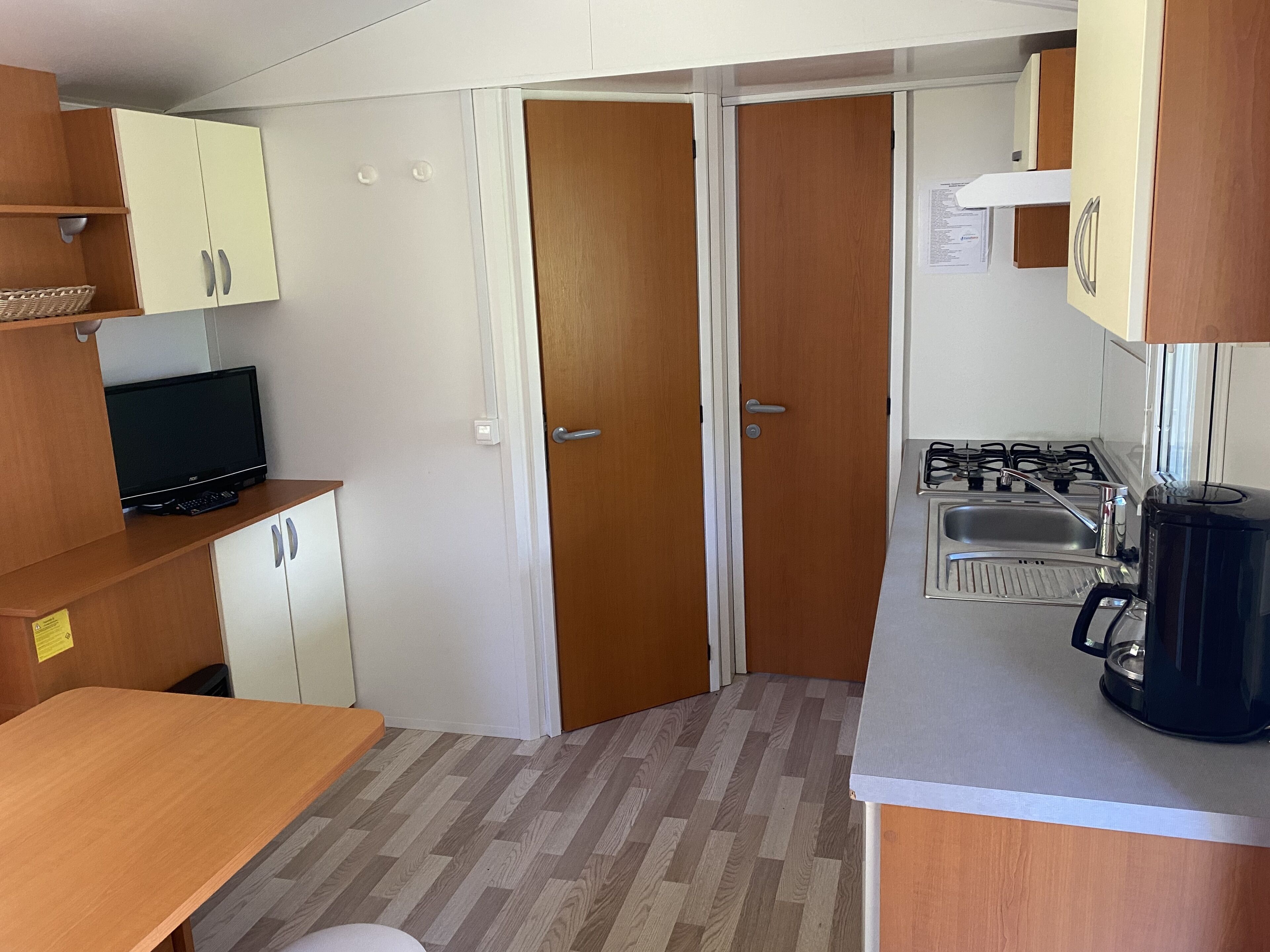 A compact kitchen with a stove, sink, and a coffee maker. There is a television on a wooden shelf and a small table.
