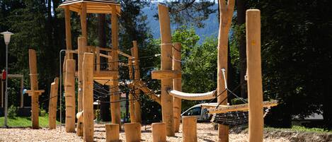 Una estructura de juego de madera en un parque, con un puente de cuerda y un pequeño refugio de madera.