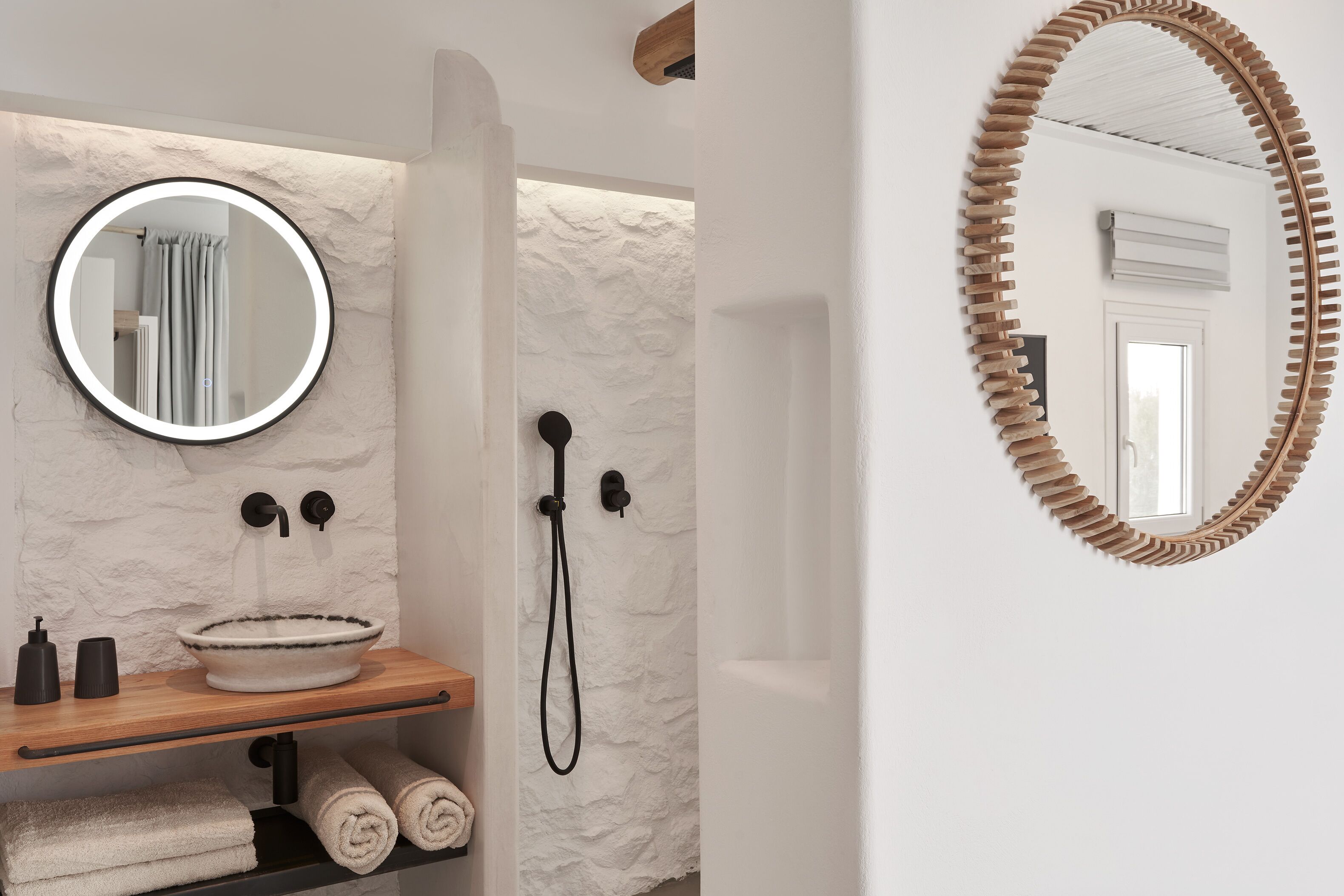 Une salle de bain avec un miroir rond, une étagère en bois avec des serviettes, un lavabo et une douche.