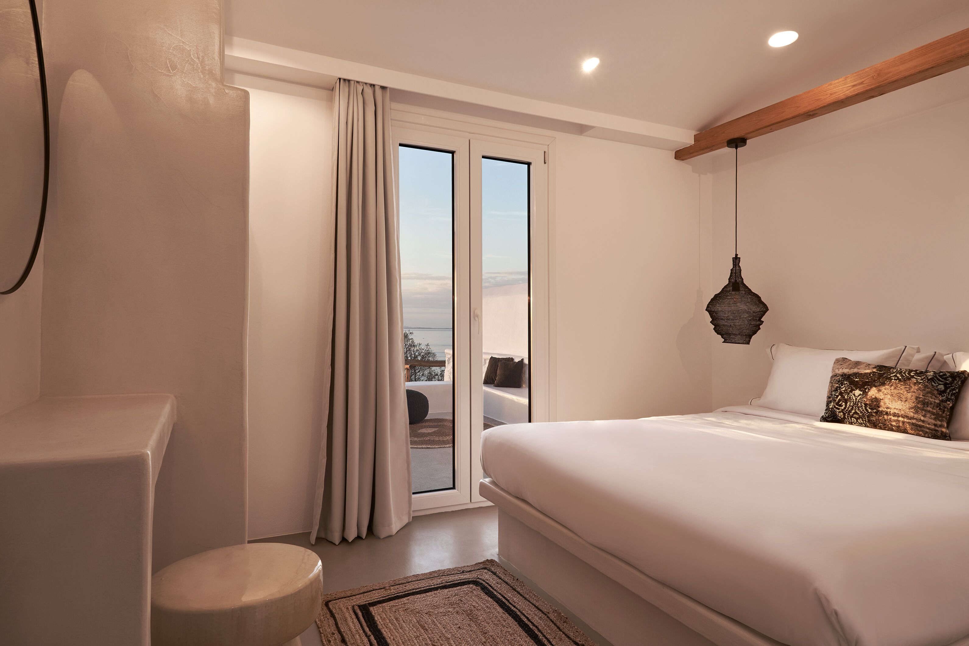 A bedroom with a large bed, a window with curtains, a hanging light fixture, and a view of the outdoors.