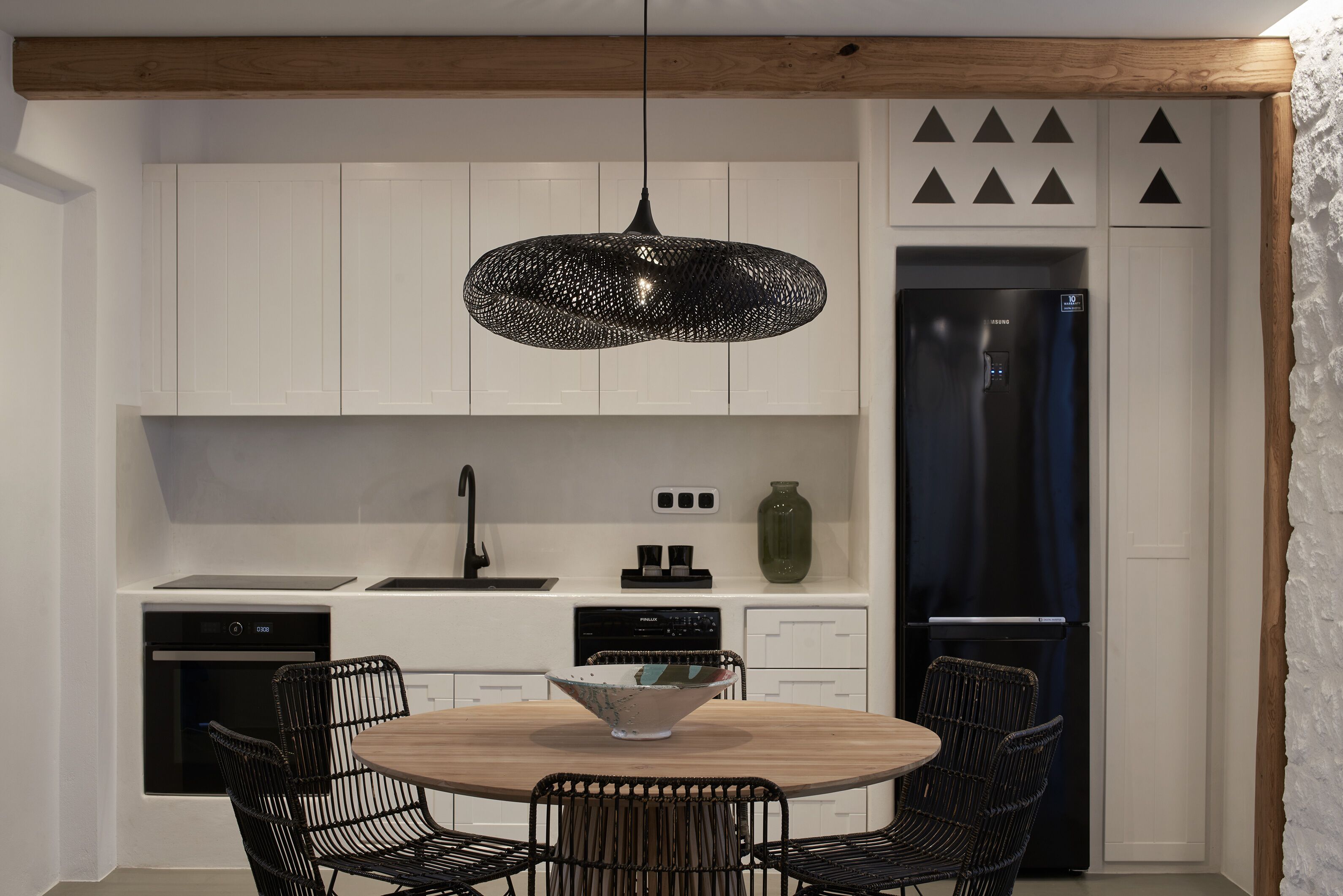 Une cuisine moderne avec des meubles blancs, un réfrigérateur noir, une table à manger en bois avec des chaises, et un luminaire suspendu noir original.