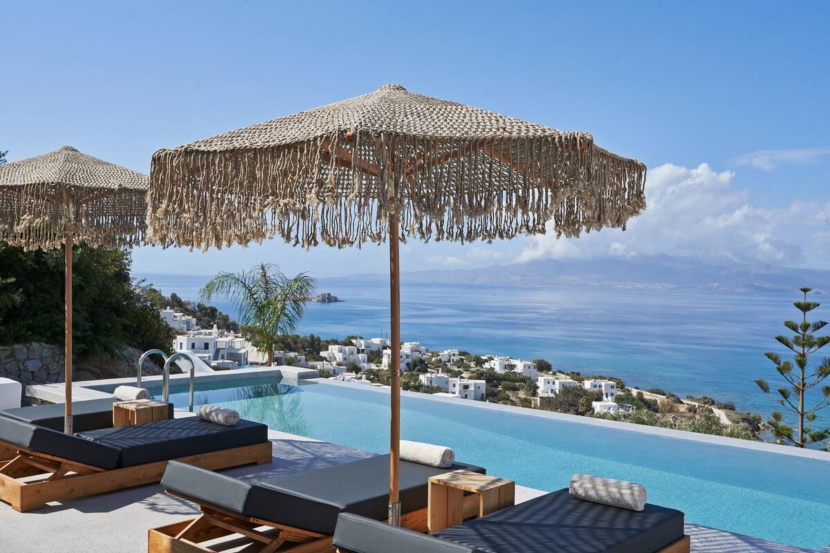 An infinity pool with lounge chairs and umbrellas, overlooking a coastal town and the sea.