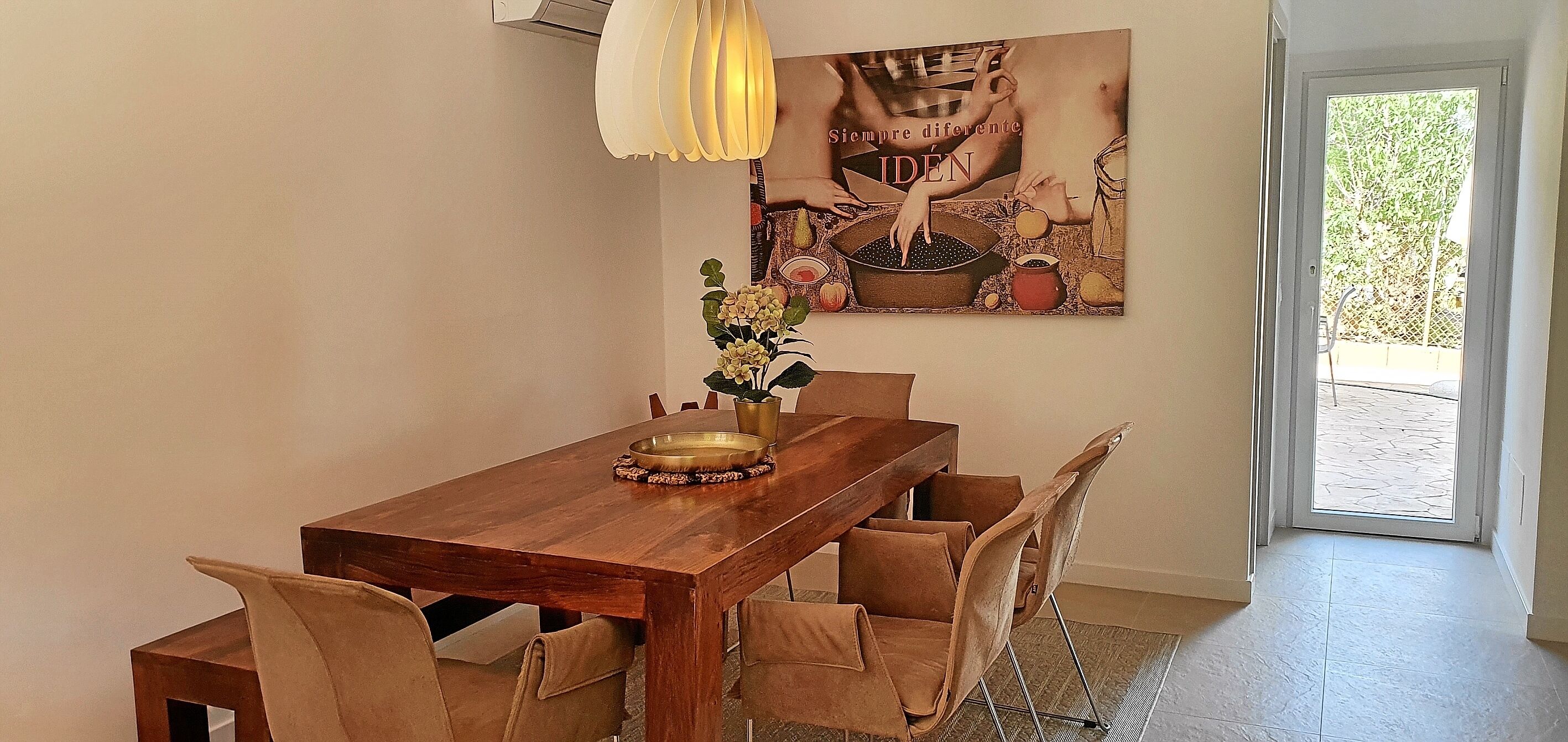 Un comedor con una mesa de madera, seis sillas, un florero con flores y un cuadro en la pared.