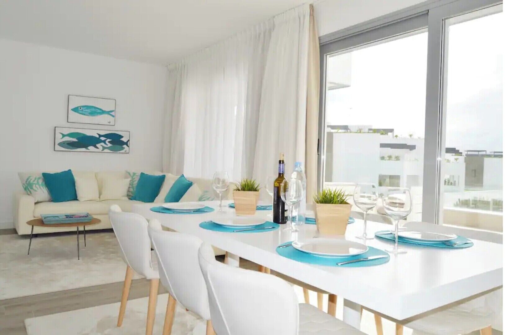 Una sala de estar moderna con comedor, que incluye un sofá con cojines turquesa, una mesa blanca lista para comer y un ventanal con vistas a la ciudad.