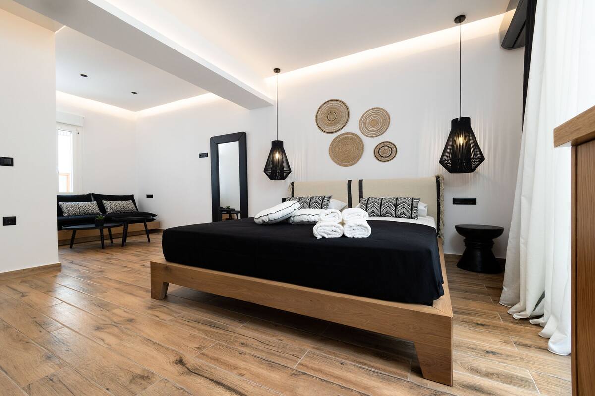 Ein modernes Schlafzimmer mit einem hölzernen Bett, einem schwarzen Kopfteil und einer schwarzen Tagesdecke. Es gibt zwei schwarze Pendelleuchten, einen Spiegel und Wandkunst aus Gewebe.