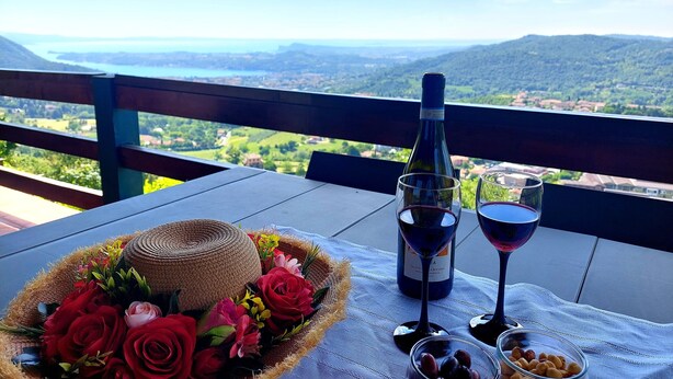 A table with a bottle of wine, two wine glasses, a bowl of olives, and a basket of flowers on a balcony overlooking a scenic landscape. - Bright apartment with pool and amazing view (Mezzane)