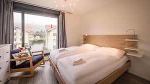 A modern bedroom with a large bed, a bedside table, a chair, and a view of buildings through a window. - Central luxury apartment with stunning views, lift access and wellness area (Matten bei Interlaken)