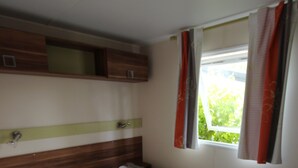 Een enkel bed met een witte matras en een blauw-oranje deken, een houten hoofdbord en een raam met gordijnen.
