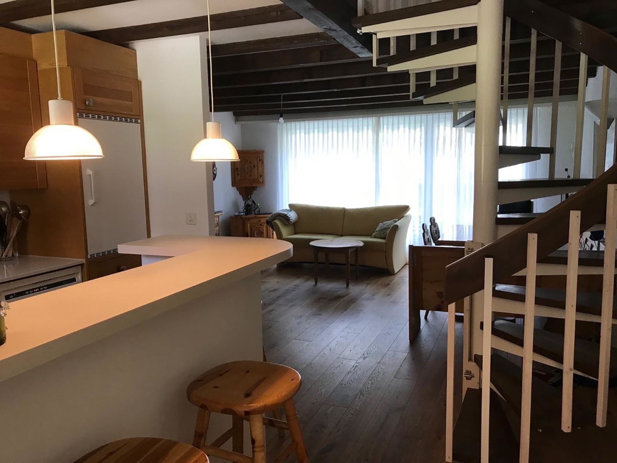 A modern kitchen with a wooden counter and stools, a dining area with a sofa and table, and a staircase with a white railing.
