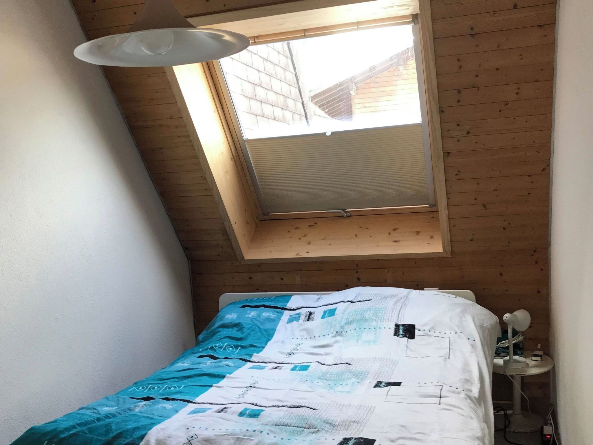 A bedroom with a sloped ceiling, a skylight, a bed with a patterned bedspread, and a bedside table with a lamp.