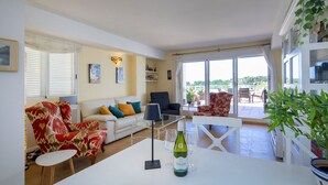 A spacious living area with a white sofa, red patterned armchairs, a glass coffee table, and a dining table with a bottle of wine.