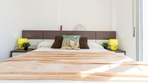 Un lit bien fait, recouvert d’une couverture beige, avec deux tables de chevet dotées de lampes et une tête de lit.