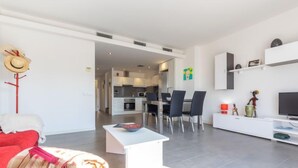 Un salon moderne avec un canapé, un coin repas avec des chaises, une cuisine en arrière-plan et un téléviseur posé sur un meuble.