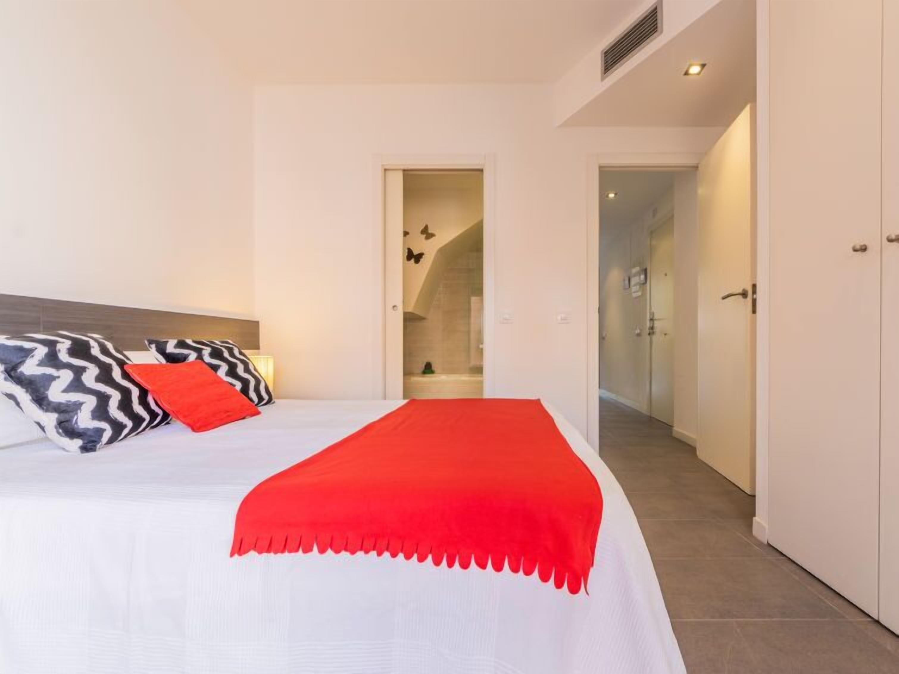 Ein Schlafzimmer mit einem Bett, einer roten Tagesdecke und einem schwarz-weißen Kissen. Eine Tür führt zu einem Badezimmer.