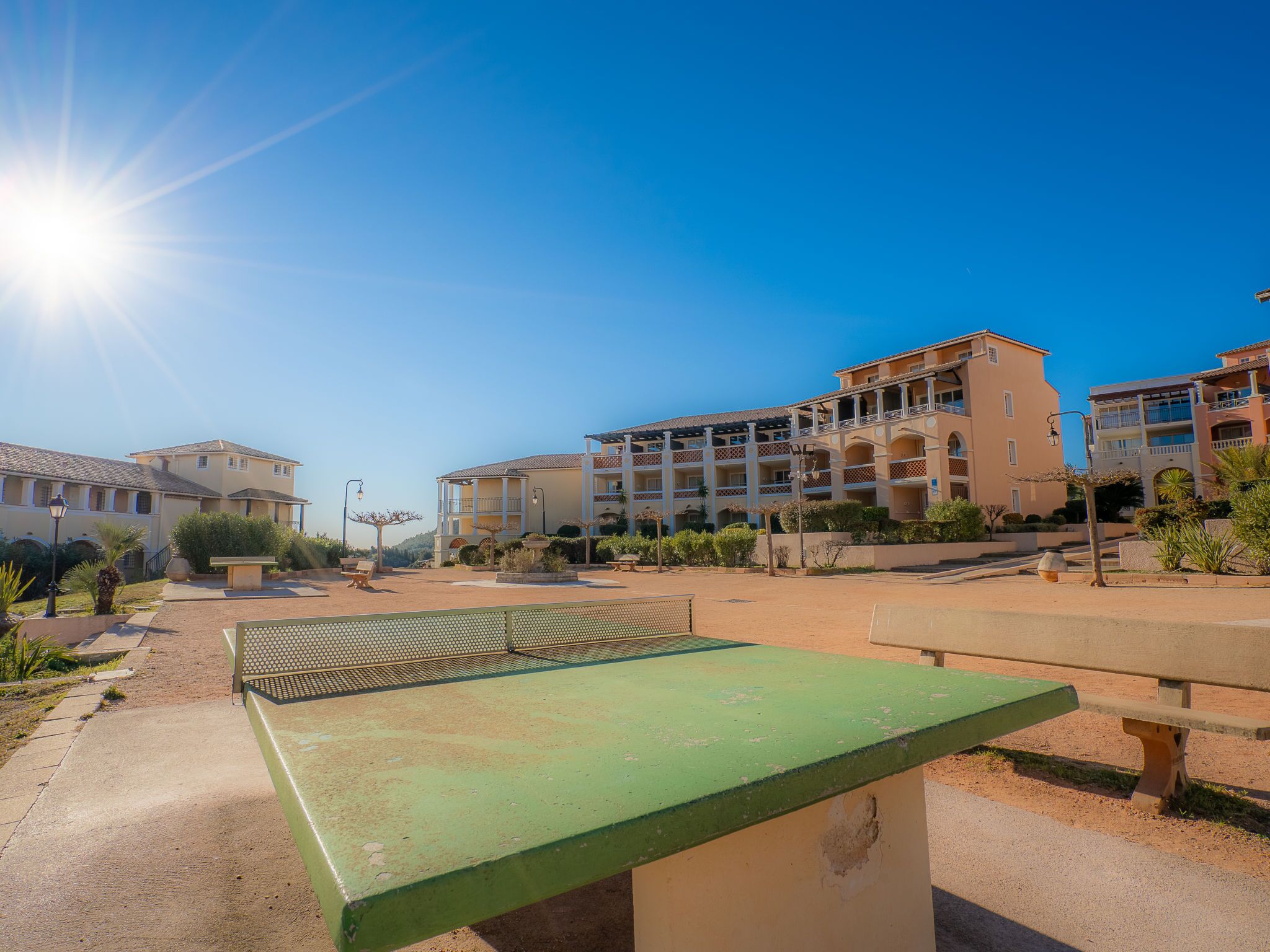 A sunny outdoor area with a green table tennis table, benches, and a building with multiple balconies and windows.