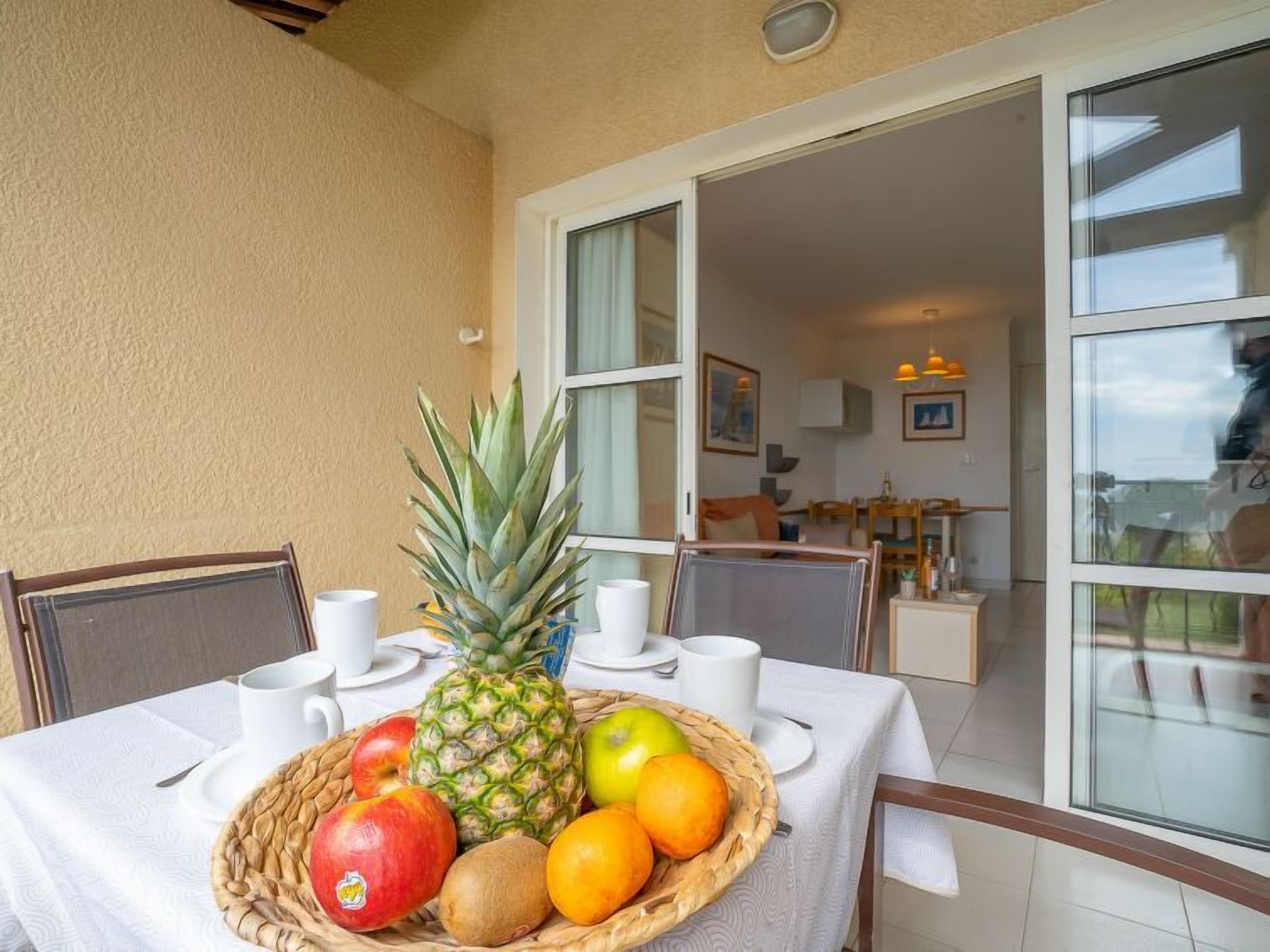 A dining area with a table set for two, featuring a fruit basket with apples, oranges, and a pineapple, and white cups and saucers.