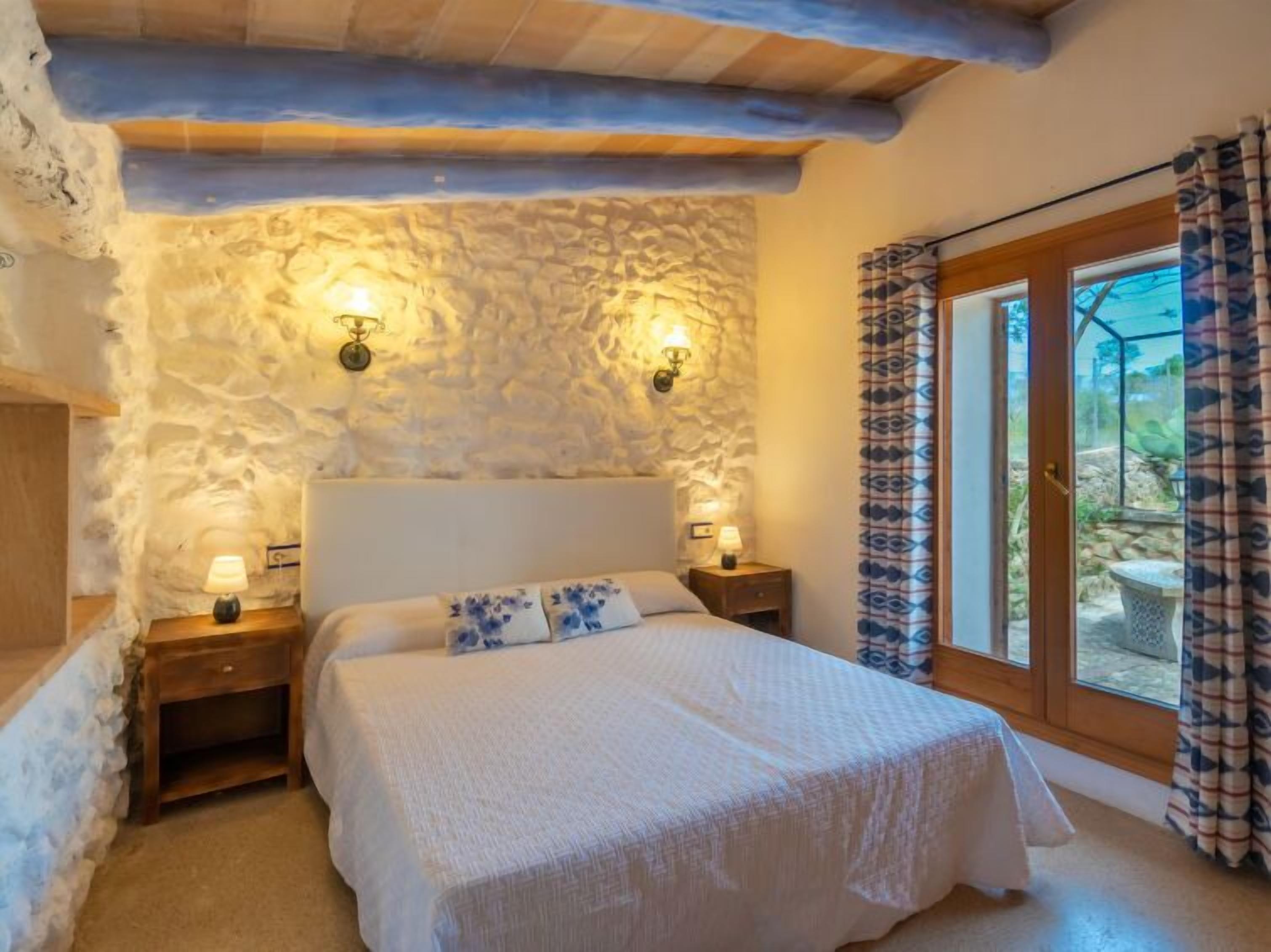 A bedroom with a stone wall, a bed with a white bedspread, two bedside lamps, and a view through a glass door.