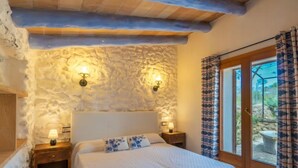 A bedroom with a stone wall, a bed with a white bedspread, two bedside lamps, and a view through a glass door. - Capella Rubi by Interhome (Artà)