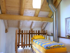 Interior de una cabaña de madera con una cama, un tubo de metal y una ventana con cortina de encaje.