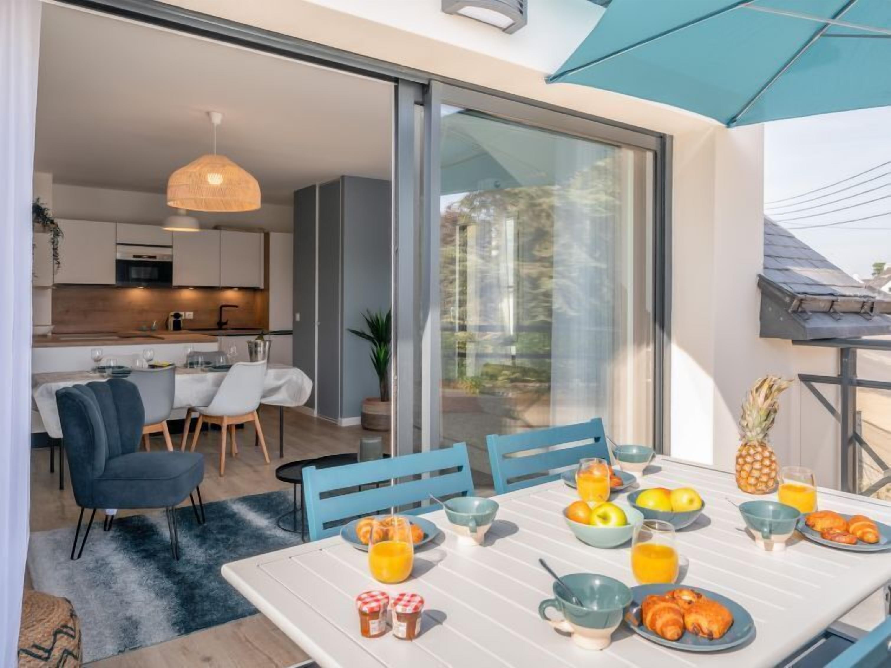 Un espace repas moderne, avec une table mise pour le petit-déjeuner, comprenant des fruits et des boissons, et offrant une vue sur la cuisine et l’espace extérieur.