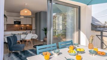 Un espace repas moderne, avec une table mise pour le petit-déjeuner, comprenant des fruits et des boissons, et offrant une vue sur la cuisine et l’espace extérieur.