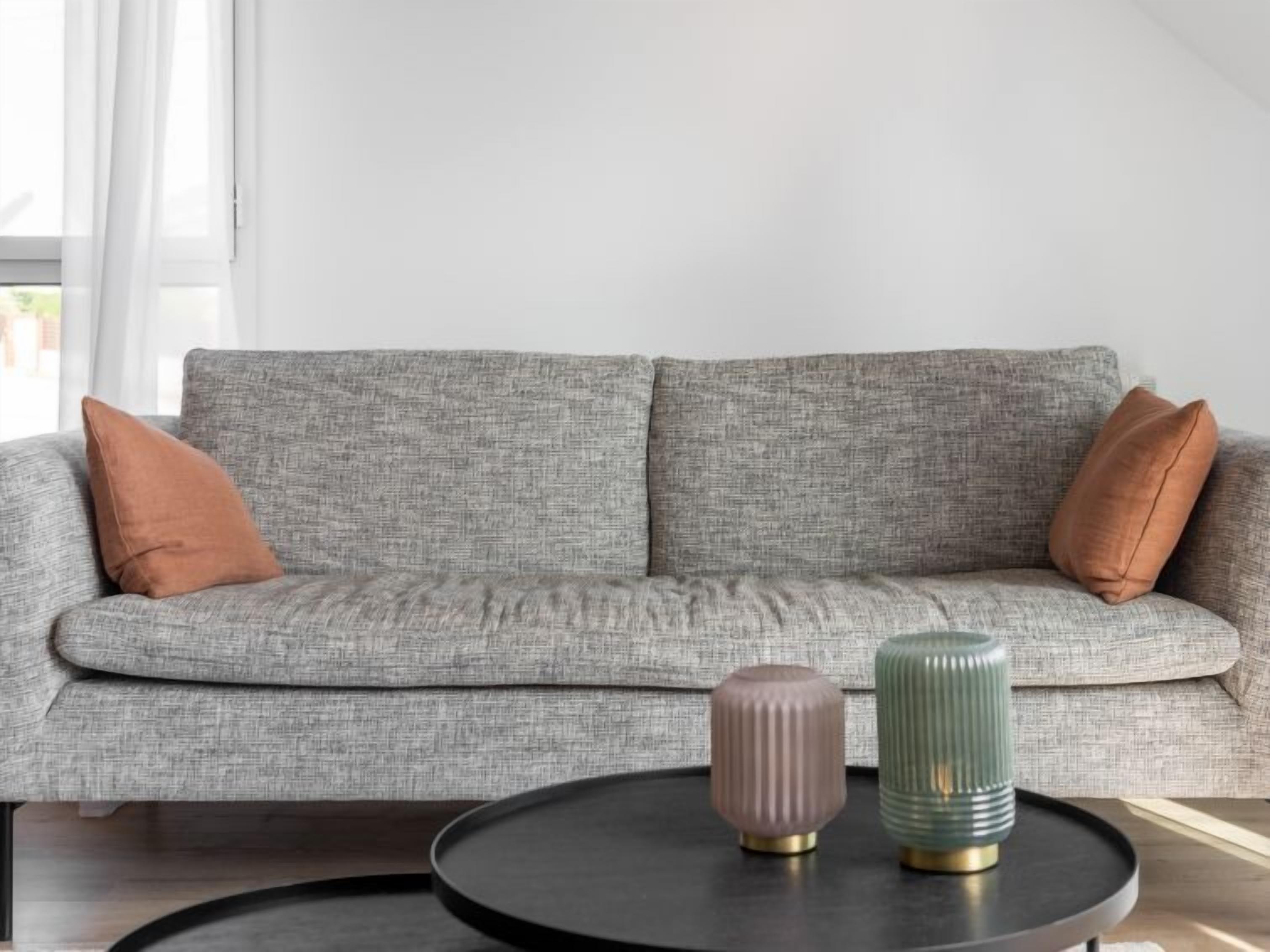 Un canapé gris avec deux coussins marron, une table ronde noire ornée de deux vases décoratifs et un mur blanc avec une fenêtre.