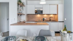 Une cuisine moderne avec des armoires blanches, un plan de travail en bois et un four encastré. On y trouve un coin repas avec une table dressée pour deux personnes, et une bouteille de vin dans un seau à glace.