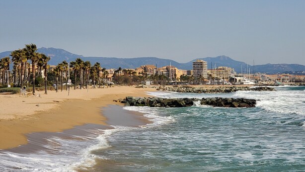 A coastal beach with a rocky breakwater, palm trees, and buildings in the background. - Port FREJUS Studio with sleeping area of ​​30 m2 for 2 Adults 2 Children with quiet balcony on the garden side (Fréjus)