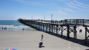 A beach with a wooden pier extending into the ocean, a colorful umbrella, and people walking on the sand. - Premier Resort: Oceanfront w/Amenities, Restaurants & Amazing Views-North Myrtle Beach (North Myrtle Beach)