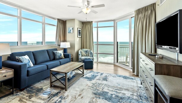 A modern living room with a sofa, coffee table, and a TV. Large windows offer a view of the ocean. - Premier Resort: Oceanfront w/Amenities, Restaurants & Amazing Views-North Myrtle Beach (North Myrtle Beach)