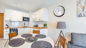 A modern kitchen with white cabinets, a microwave, and a sink. There is a dining table with placemats and chairs. A clock and a lamp are on the wall. - Apartamenty Sun & Snow Bursztynowa (Sztutowo)