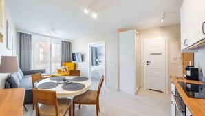 A modern apartment with a dining area, a living room with a sofa and a yellow armchair, a kitchen with a stove and a coffee machine, and a bedroom visible through an open door. - Apartamenty Sun & Snow Bursztynowa (Sztutowo)