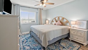 Une chambre à coucher comprenant un lit, des tables de chevet, un ventilateur de plafond, une fenêtre avec des stores et un tapis à motifs.