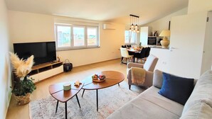 A modern living room with a flat-screen TV, a dining area with a table and chairs, and a kitchen in the background.