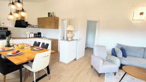 A modern living room with a grey sofa, a dining area with a wooden table and chairs, and a kitchen area with wooden cabinets and a coffee machine.