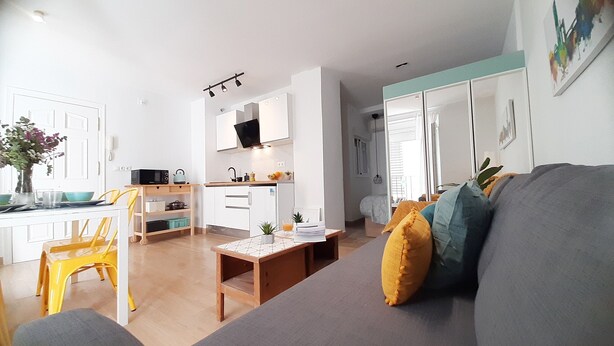 A compact living space with a kitchenette, a dining area with a yellow chair, and a grey sofa with cushions. - Lovely penthouse apartment with terrace Lovely Center Apartments Alameda (Sevilla)