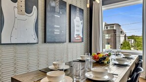 Un comedor con una larga mesa de madera dispuesta para cuatro personas, adornado con ilustraciones de guitarras en la pared y vistas de edificios al exterior.