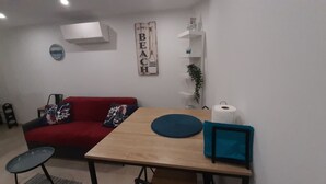 Un salon avec un canapé rouge, une table en bois, une petite table ronde, un climatiseur fixé au mur, une étagère avec des objets décoratifs et une décoration murale sur laquelle est inscrit le mot « PLAGE ».