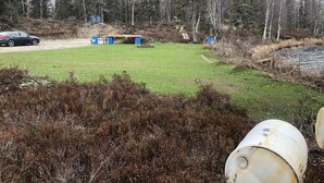 A grassy field with a car and barrels, surrounded by trees. - Private Lake Front with boat launch and dock. (Kenai)