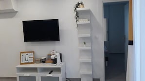 Una habitación con un televisor de pantalla plana montado en la pared, un escritorio pequeño con una cafetera, un letrero que dice "10 AOU" y una planta en un estante.